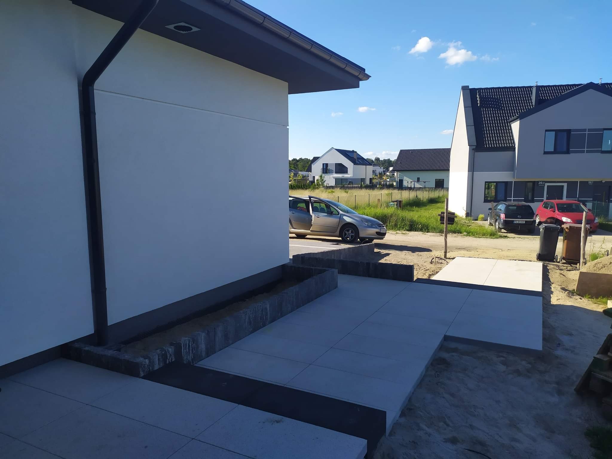 Nowoczesny taras z jasnoszarych płyt betonowych w trakcie budowy, widoczne krawężniki, rynna i fragment elewacji budynku, w tle osiedle domów jednorodzinnych i samochody.