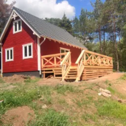 Czerwony dom z drewnianym tarasem i schodami, widok z dołu, częściowo zarośnięty teren, błękitne niebo z obłokami.