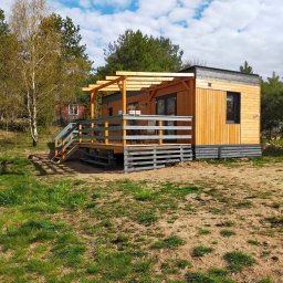 Luxdrew Robert Wenta - Nowoczesny drewniany domek letniskowy z tarasem i pergolą, wykończony w jasnym drewnie i szarościach, otoczony zielenią i piaszczystym podłożem.