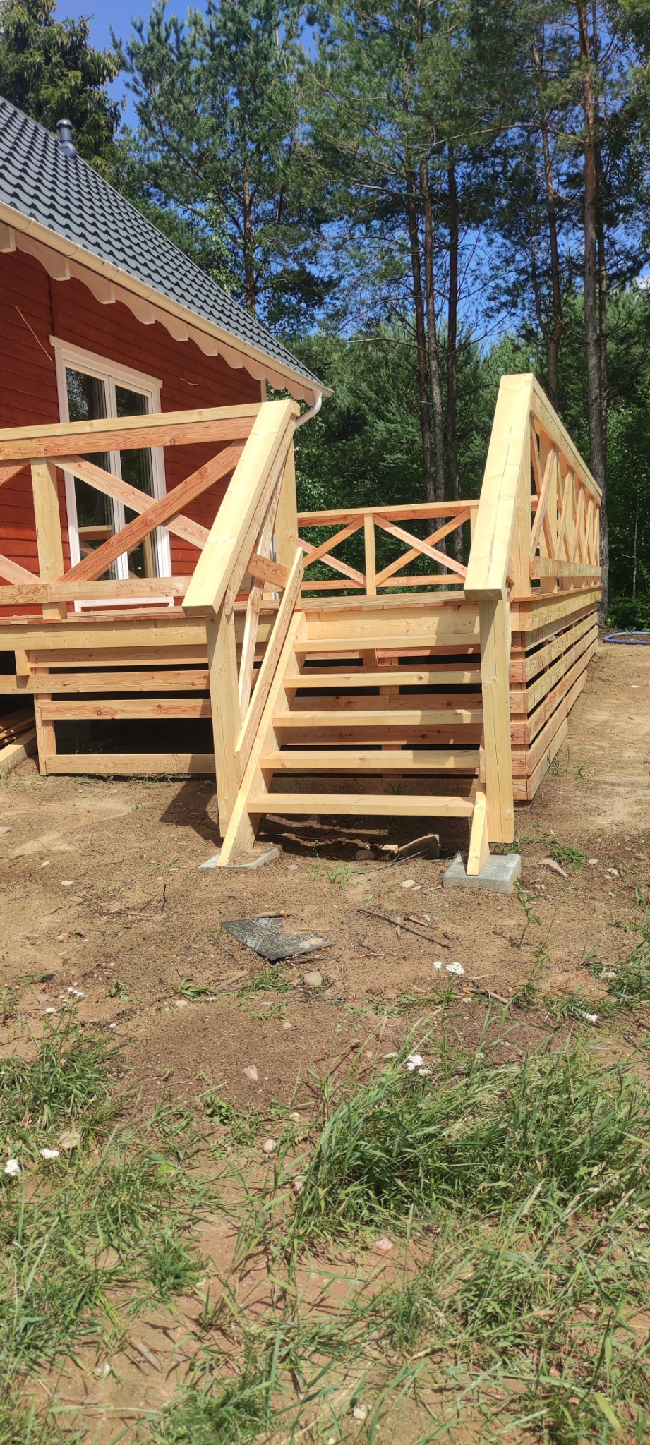 Surowy, drewniany taras z balustradą i schodami, w trakcie budowy, z widocznym fragmentem domu z czerwonymi ścianami i zielonym lasem w tle.