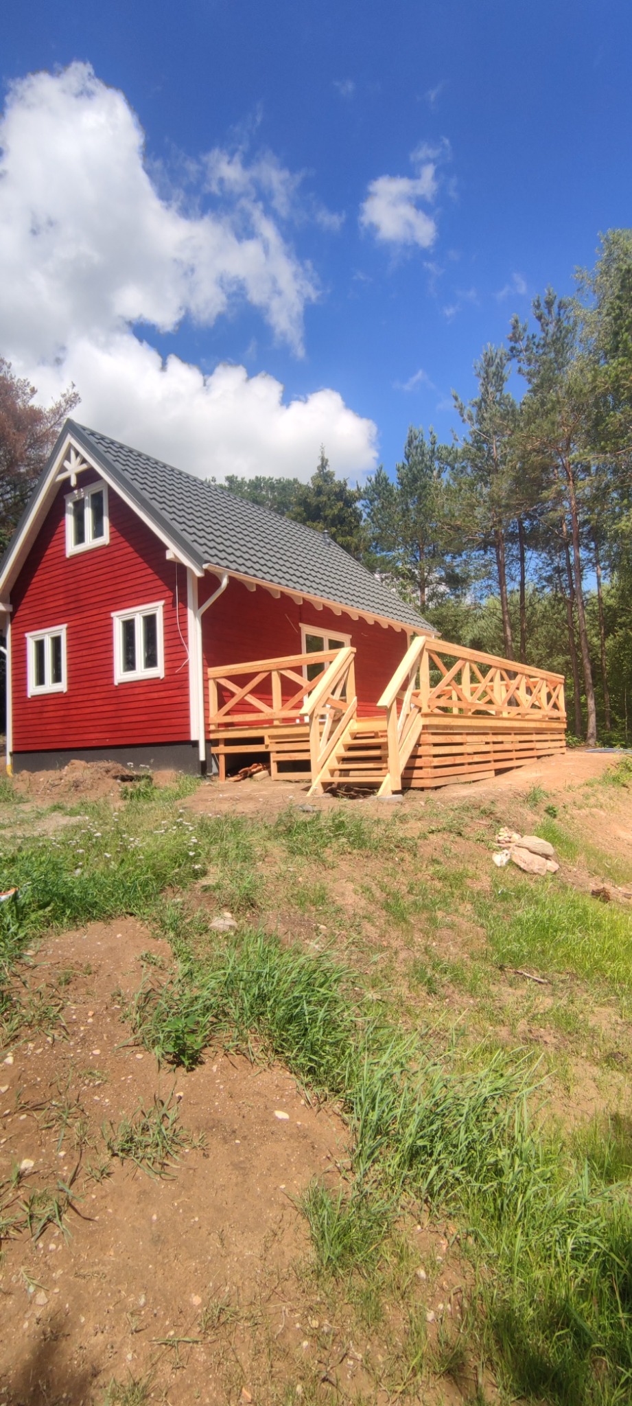 Czerwony dom z drewnianym tarasem i schodami, widok z dołu, częściowo zarośnięty teren, błękitne niebo z obłokami.
