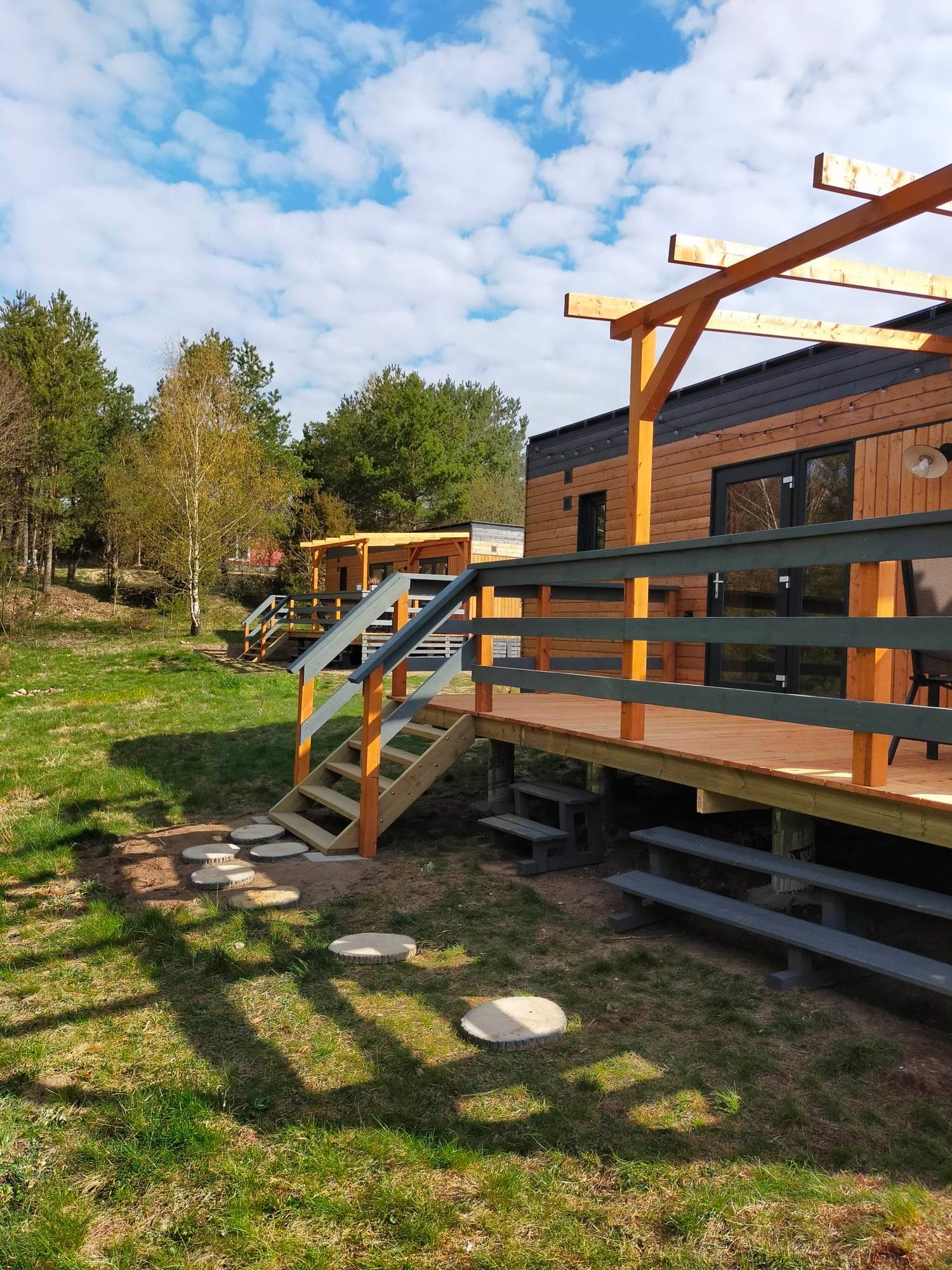 Dwa domki letniskowe z drewnianą elewacją, tarasami i pergolami, połączone ścieżką z drewnianych krążków na trawniku.