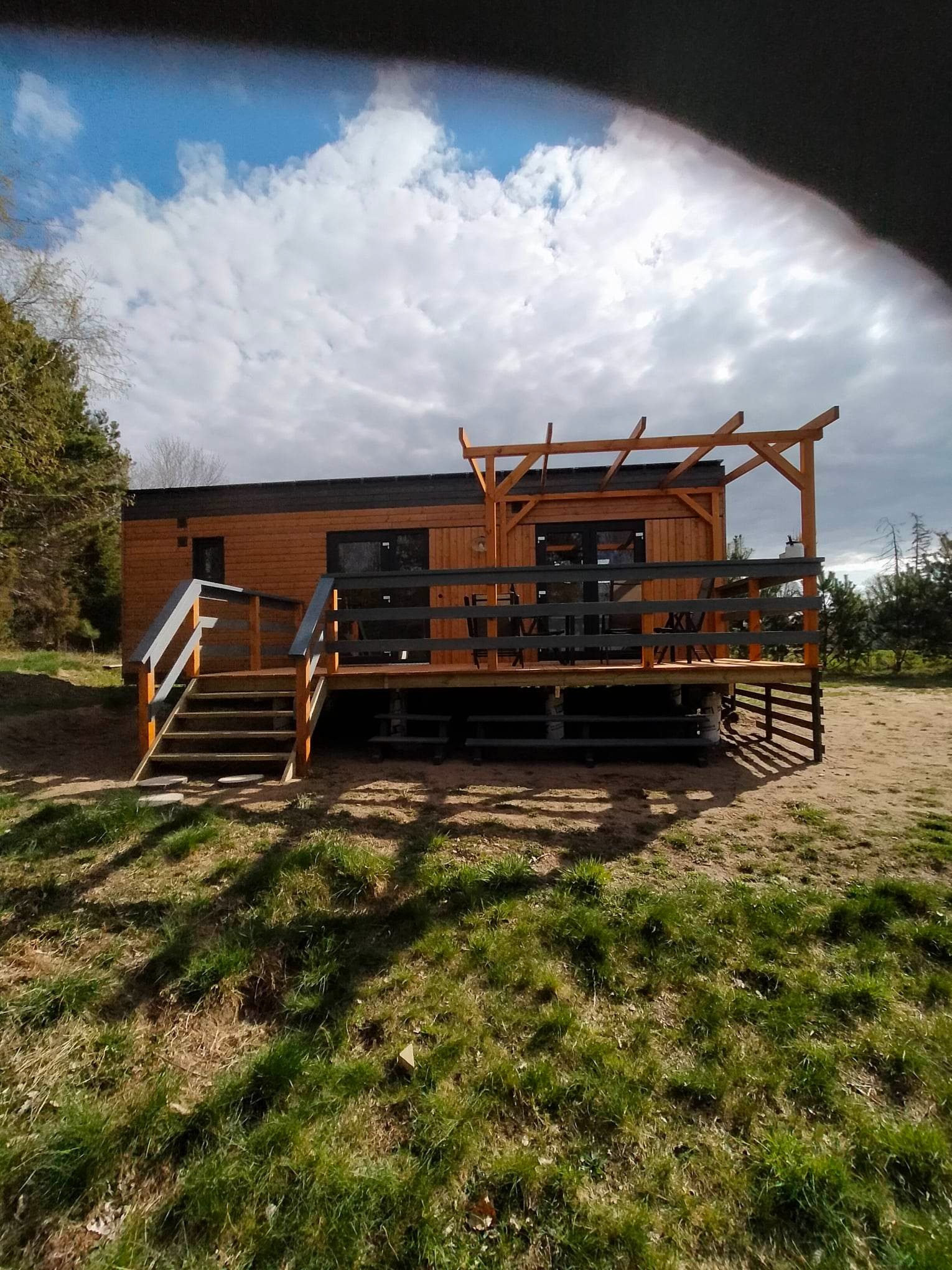 Drewniany domek z tarasem i pergolą w słoneczny dzień, widok z poziomu gruntu, zarys drzew na horyzoncie.