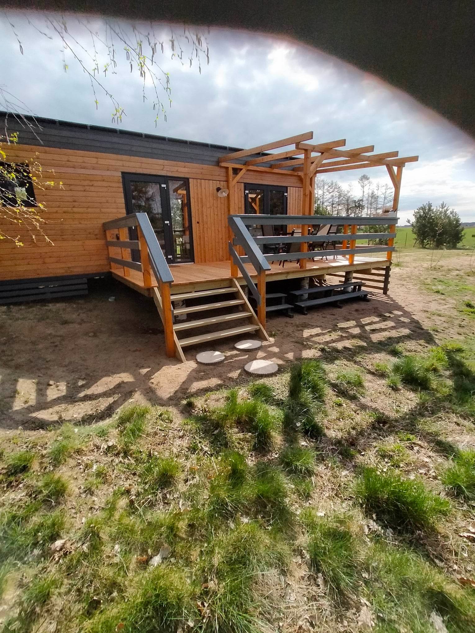 Drewniany dom z tarasem i pergolą w surowym otoczeniu, widok z poziomu trawy. Schody z okrągłymi kamieniami prowadzą na podest z szarą balustradą i ażurową pergolą.