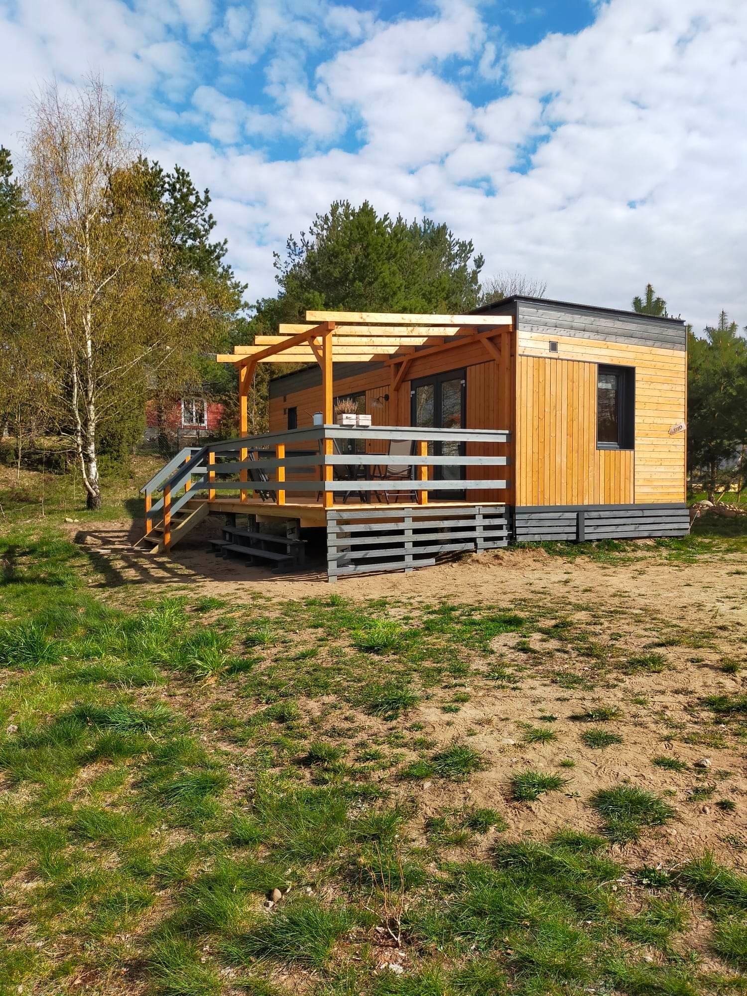 Nowoczesny drewniany domek letniskowy z tarasem i pergolą, wykończony w jasnym drewnie i szarościach, otoczony zielenią i piaszczystym podłożem.