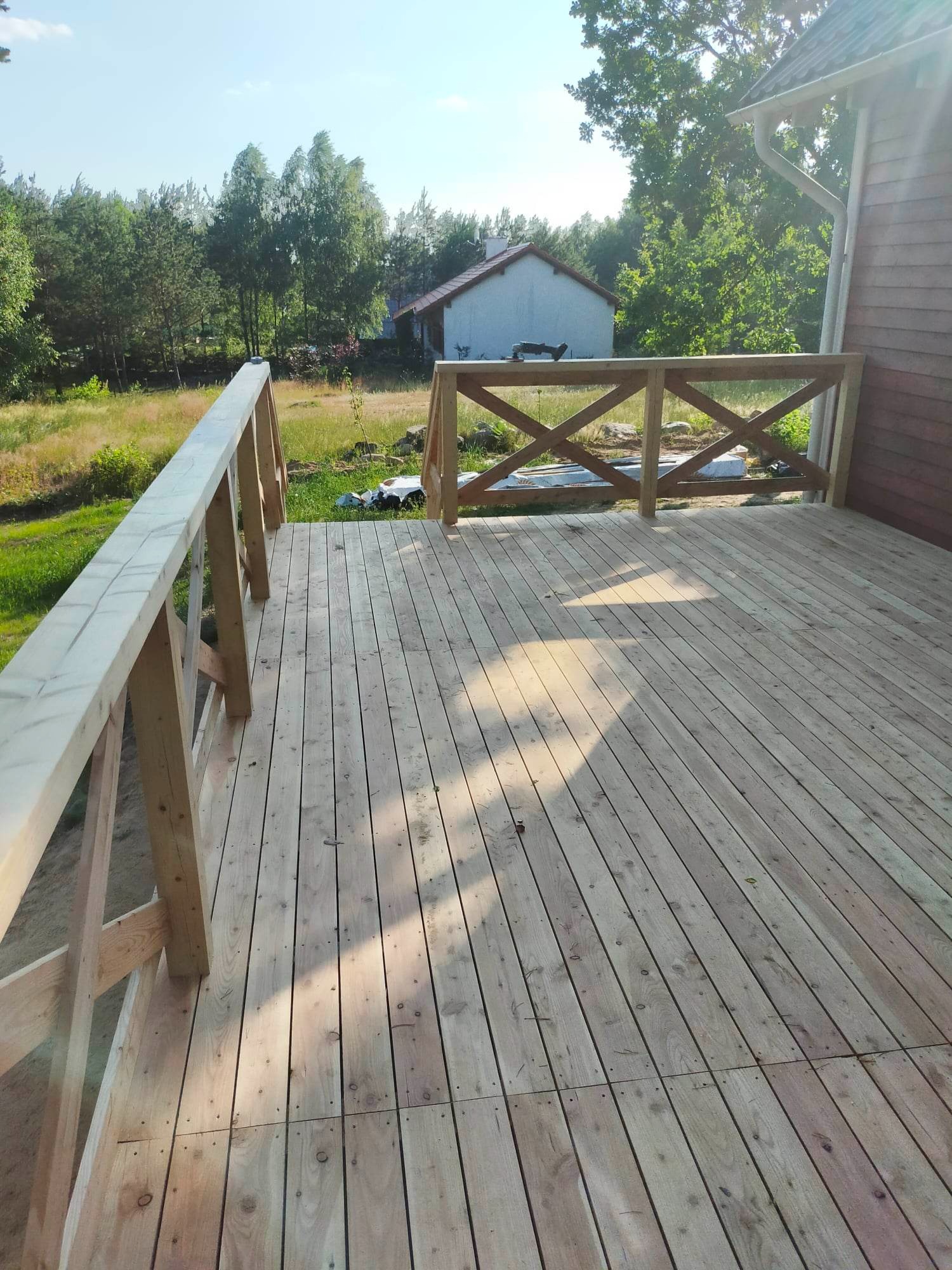 Świeżo wybudowany taras z jasnego drewna z krzyżową balustradą, widok na dom i zieleń w tle, oświetlony promieniami słońca.
