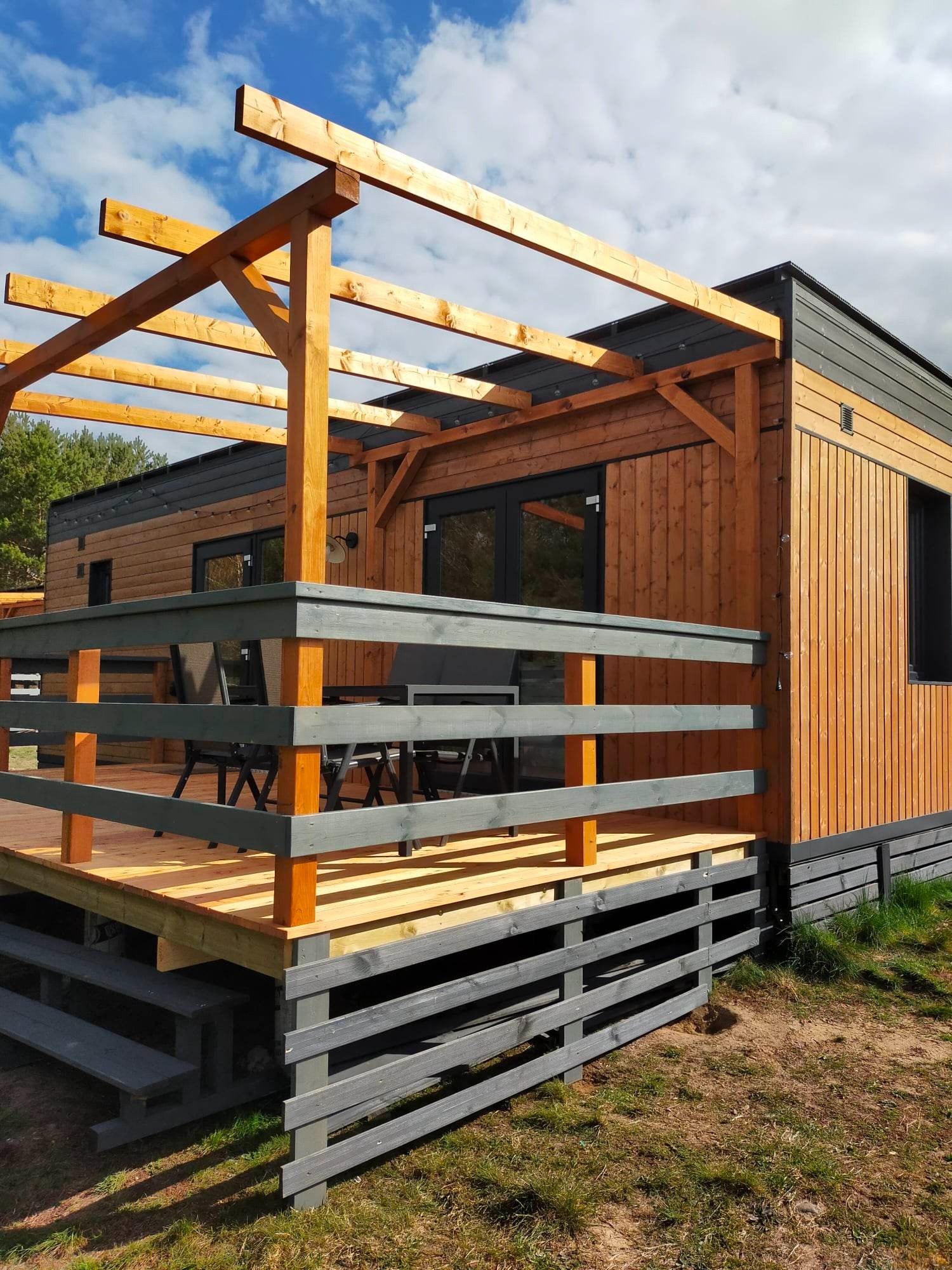 Zewnętrzny taras z drewnianą konstrukcją pergoli, szare balustrady i schody, widoczne meble ogrodowe, jasne drewno desek tarasowych, dom obłożony drewnianą boazerią w dwóch odcieniach brązu...