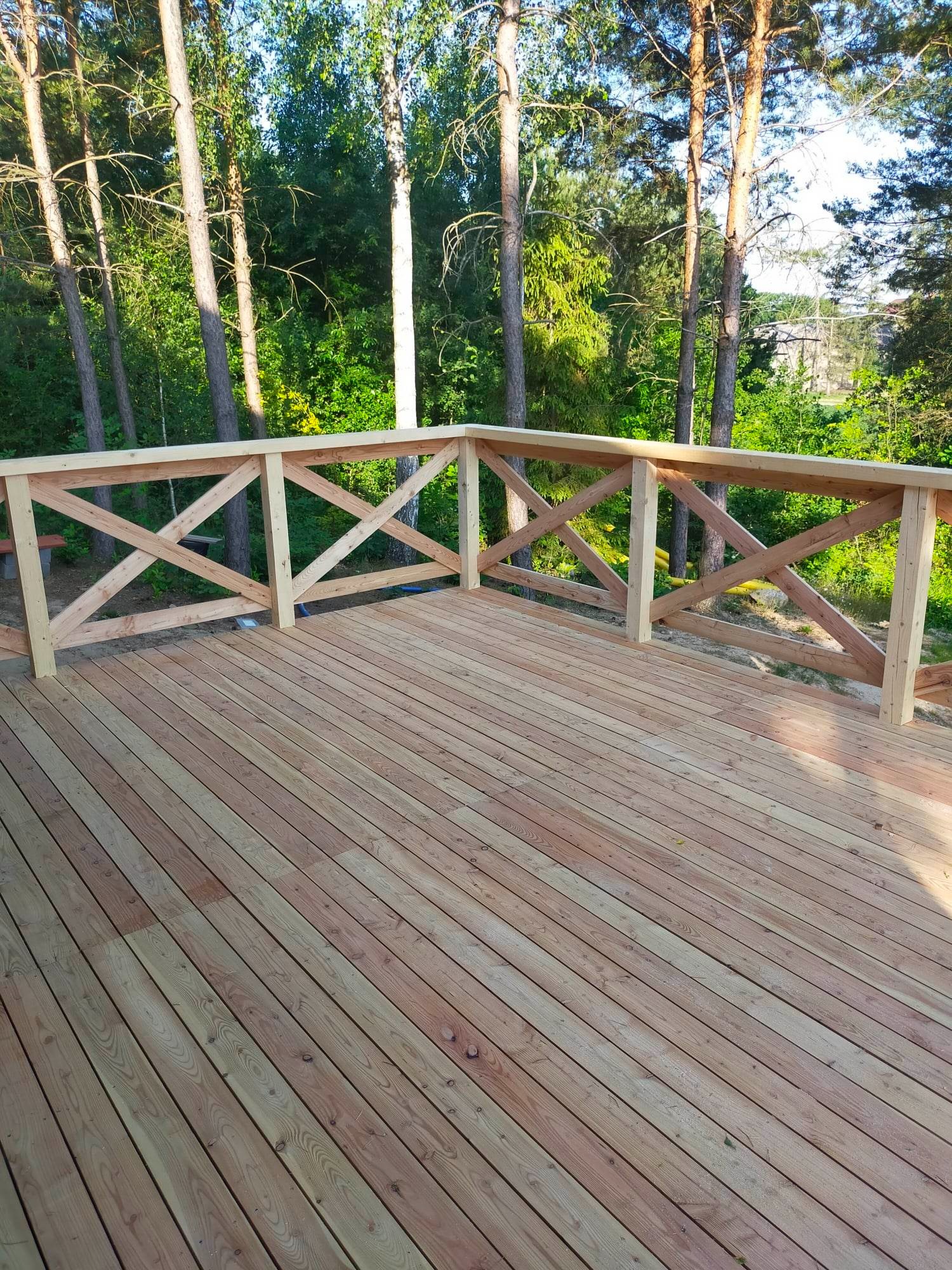 Świeżo wybudowany taras z jasnego drewna z balustradą w kształcie krzyży, otoczony zielenią drzew.
