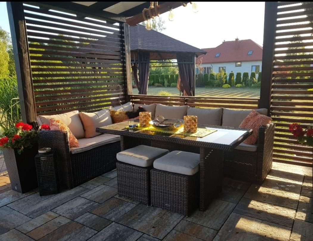 Zadaszony taras z meblami ogrodowymi z rattanu: sofa z beżowymi poduszkami, stół z blatem ze szkła i dwa pufy. Na stole dekoracje, w tle dom z czerwonym dachem i zadbany ogród.