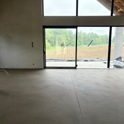 Budjan - Jan Sikora - Gładka, szara posadzka w surowym wnętrzu domu z dużymi oknami i widokiem na pole. Ściany przygotowane do tynkowania, widoczne instalacje elektryczne.