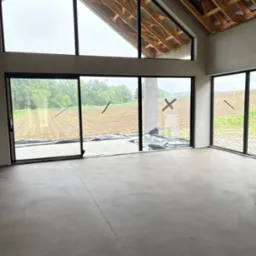 Surowe wnętrze domu z widokiem na pole przez panoramiczne okna. Szara posadzka, drewniana konstrukcja dachu. Wnętrze w trakcie wykończenia, widoczne zabezpieczenia okien.