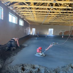 Budjan - Jan Sikora - Wyrównywanie wylewki maszyną i ręcznie w przestronnym pomieszczeniu z drewnianą konstrukcją stropu. Widoczne okna i ściany w trakcie prac wykończeniowych.