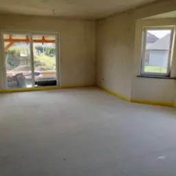 Wnętrze surowego domu w trakcie budowy, widoczne ściany przygotowane do malowania, wylana posadzka z żółtą taśmą zabezpieczającą przy ścianach oraz okna z widokiem na ogród.