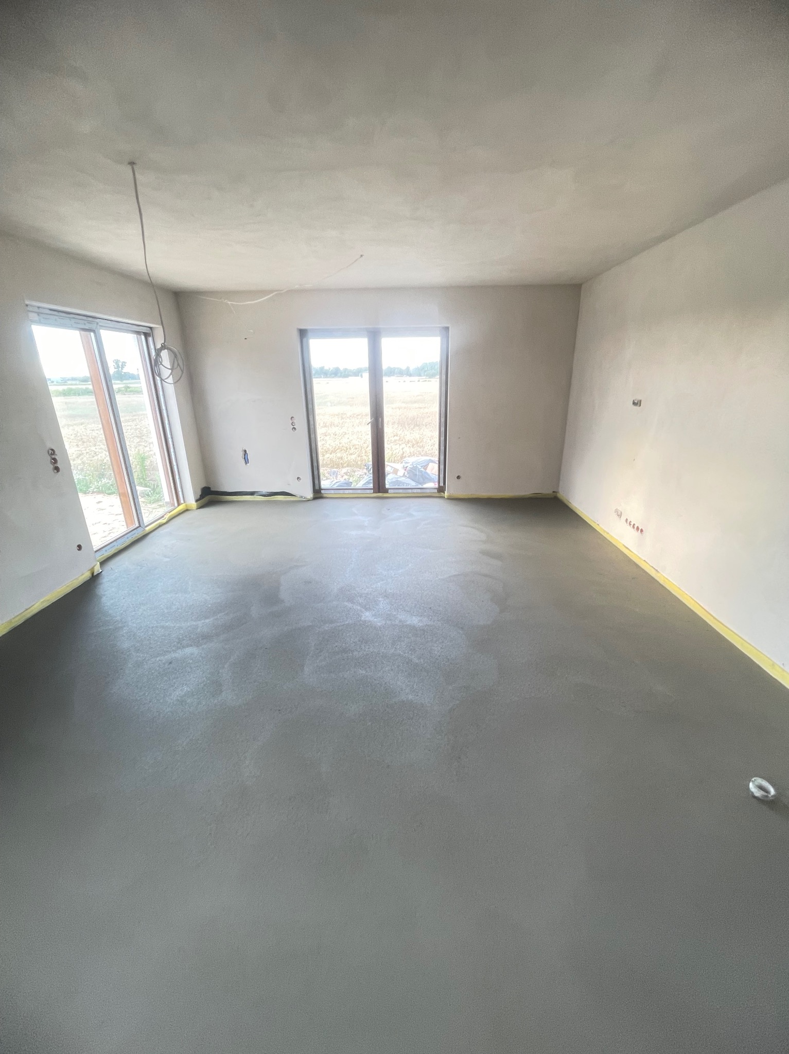 Świeżo wylana, szara posadzka w surowym wnętrzu z oknami na pole, zabezpieczona taśmą malarską przy ścianach.