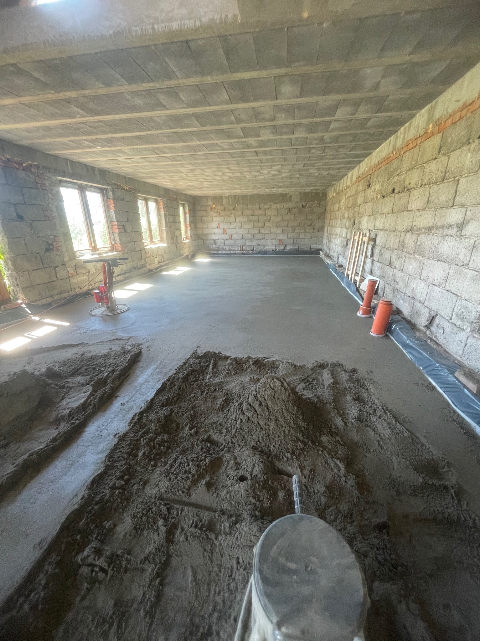 Wnętrze surowego domu w trakcie wylewania posadzki, widoczne narzędzia do rozprowadzania masy, folia izolacyjna przy ścianie oraz fragmenty cegieł w ścianach.