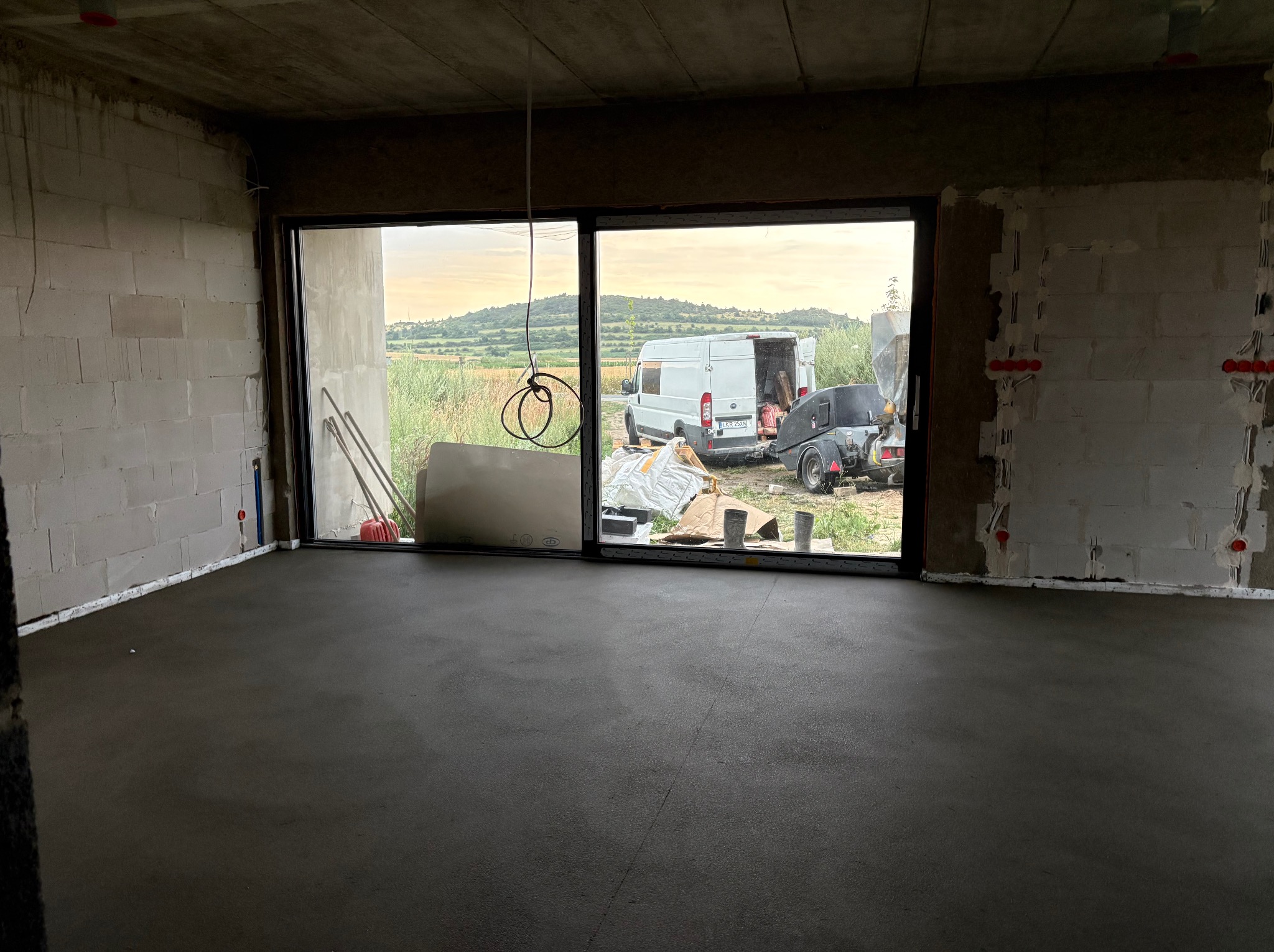 Świeżo wylana, gładka posadzka w surowym wnętrzu z widokiem na krajobraz przez duże okno. Na zewnątrz zaparkowany bus i przyczepa z betoniarką.