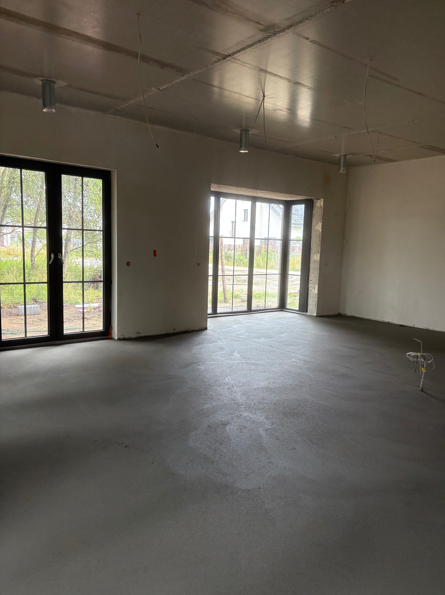 Szara, gładka wylewka w surowym wnętrzu z dużymi oknami. Widoczne instalacje pod sufitem i na podłodze. Minimalistyczny, nowoczesny styl.