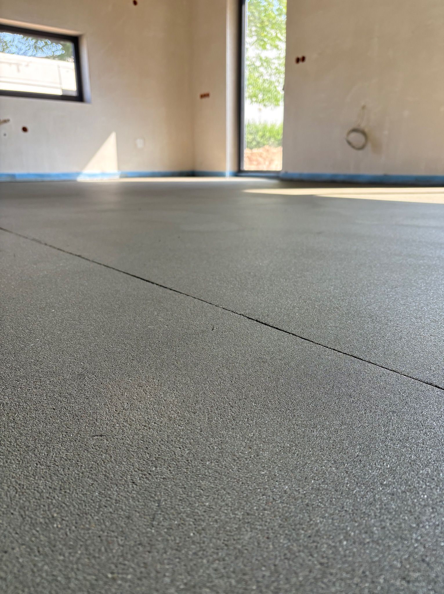 Szara, gładka posadzka w jasnym wnętrzu z oknem i przygotowanymi ścianami. Widoczna dylatacja. Minimalistyczny styl, naturalne światło.