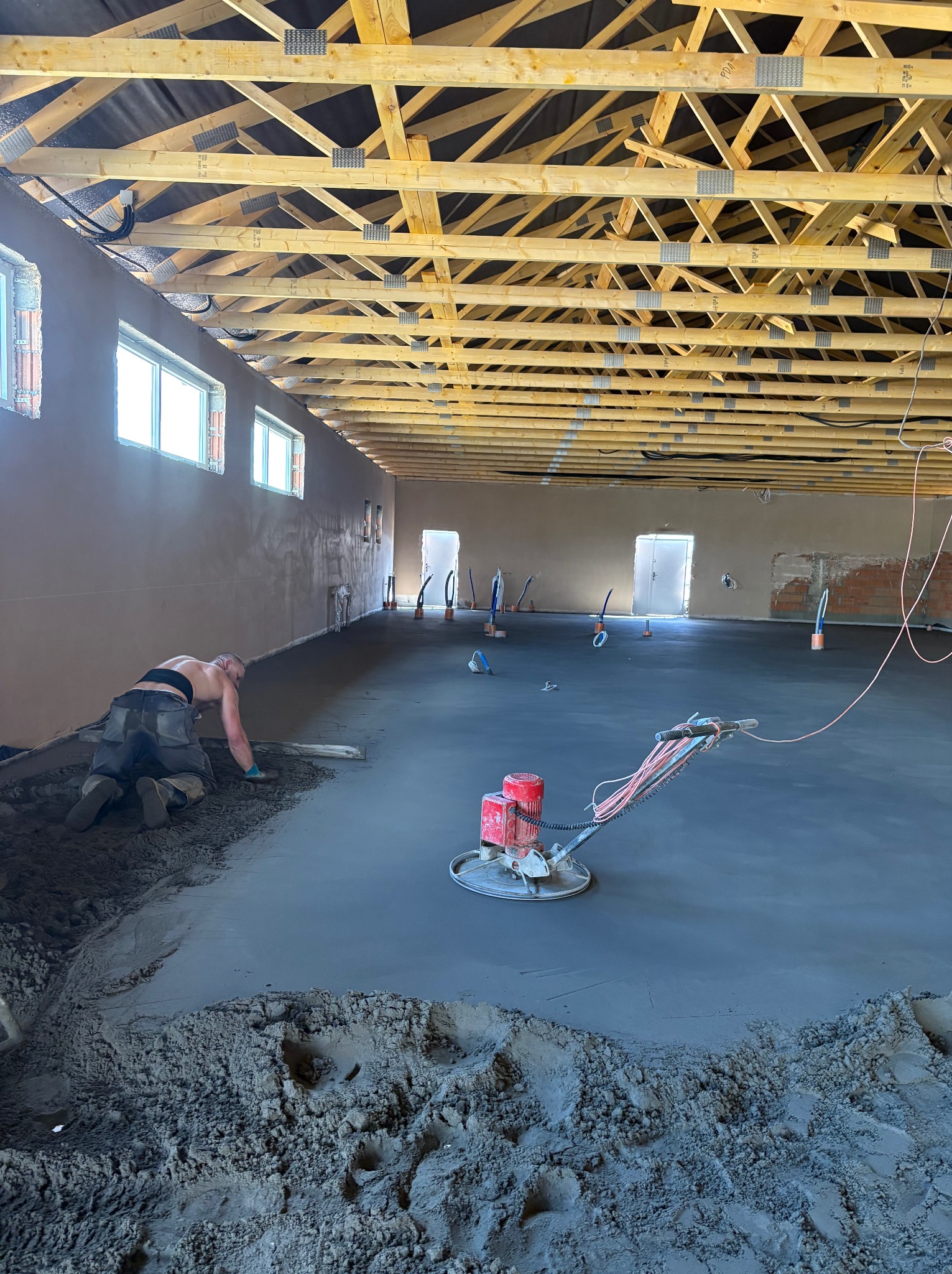 Wyrównywanie wylewki maszyną i ręcznie w przestronnym pomieszczeniu z drewnianą konstrukcją stropu. Widoczne okna i ściany w trakcie prac wykończeniowych.