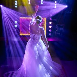 RAFAŁ GŁOGOWSKI - Panna młoda w białej sukni z koronkowym gorsetem, tyłem do obiektywu, tańczy na parkiecie w fioletowych światłach dyskoteki. DJ w tle.