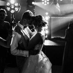 RAFAŁ GŁOGOWSKI - Czarno-białe zdjęcie tańczącej pary młodej w Szczecinie, oświetlonej reflektorami, z gośćmi weselnymi w tle. Suknia panny młodej zdobiona kwiatowym wzorem.