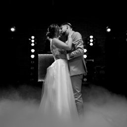 RAFAŁ GŁOGOWSKI - Czarno-białe zdjęcie całującej się pary młodej na tle świateł i dymu. Panna młoda w sukni z koronkową górą, pan młody w garniturze. Romantyczna atmosfera.