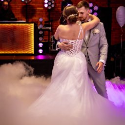 RAFAŁ GŁOGOWSKI - Para młoda w tańcu na tle ceglanej ściany z oświetleniem scenicznym i efektem mgły. Suknia z koronkowym gorsetem, garnitur w odcieniach szarości. Romantyczna atmosfera.