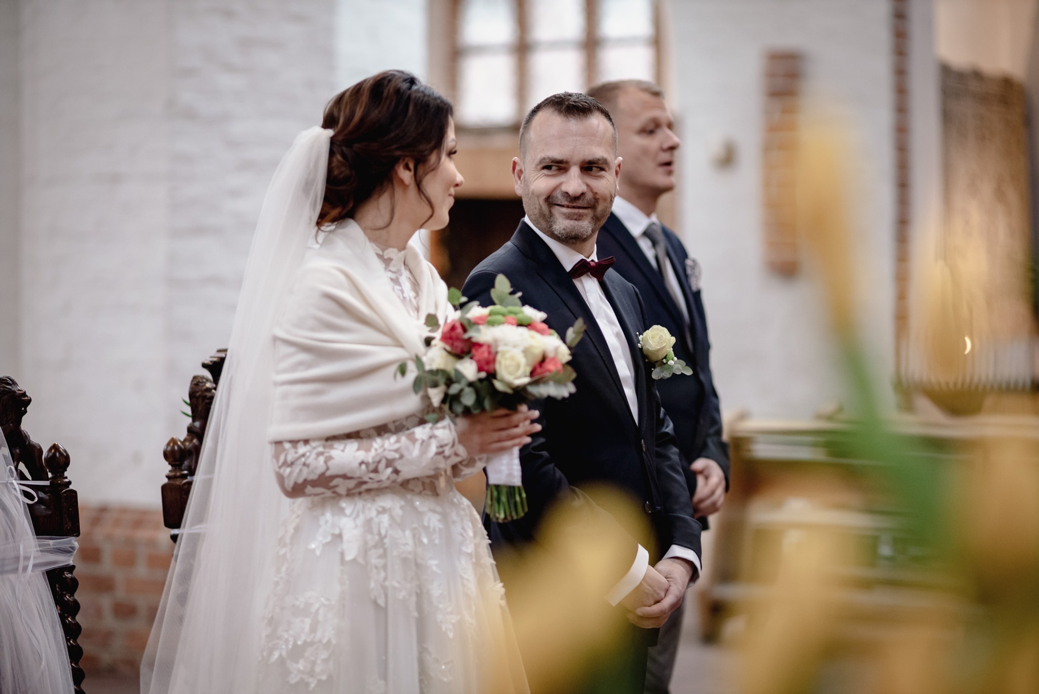 Panna młoda w koronkowej sukni i z bukietem, obok pan młody w garniturze z bordową muchą, w tle świadek w kościele.