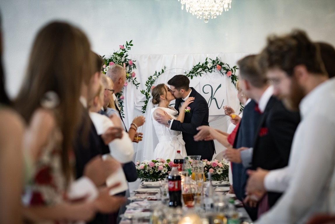 Pocałunek nowożeńców za stołem weselnym, widziany zza ramion gości, z dekoracją kwiatową i inicjałem 'K' w tle.