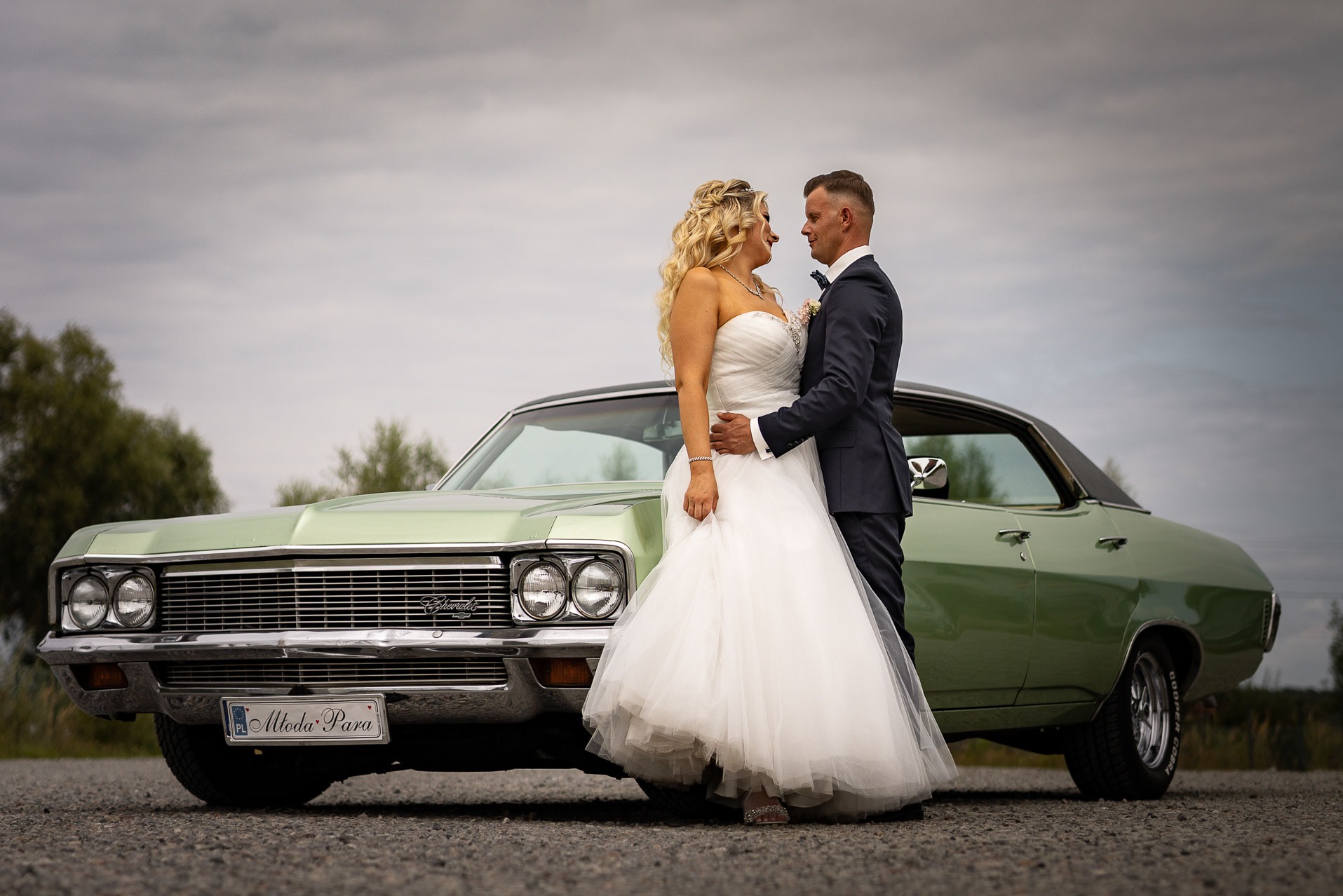 Para młoda w eleganckich strojach stoi przy odrestaurowanym, zielonym samochodzie Chevrolet Impala z tablicą rejestracyjną 'Młoda Para' na tle pochmurnego nieba.