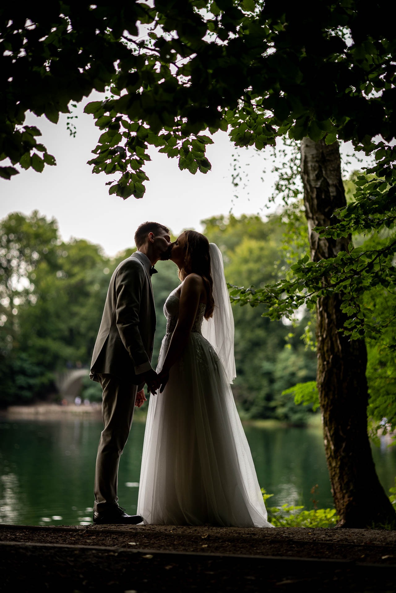 Silueta całującej się pary młodej na tle jeziora i zieleni, uchwycona spod korony drzewa. Romantyczna sceneria, stonowana kolorystyka, naturalne światło.