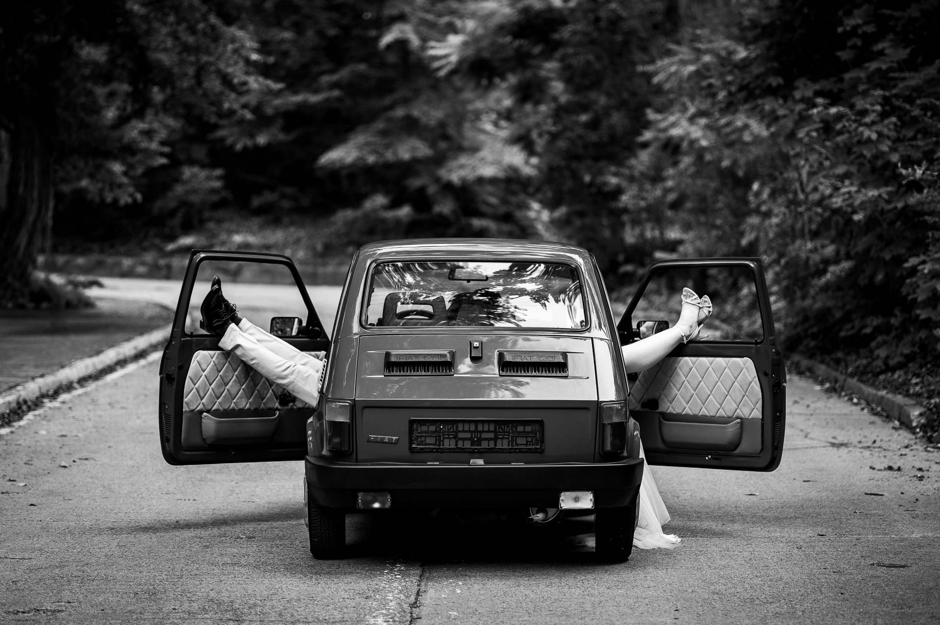 Czarno-białe zdjęcie Fiata 126p z otwartymi drzwiami, z których wystają nogi pary młodej. Nietypowe ujęcie z tyłu, na tle leśnej drogi.