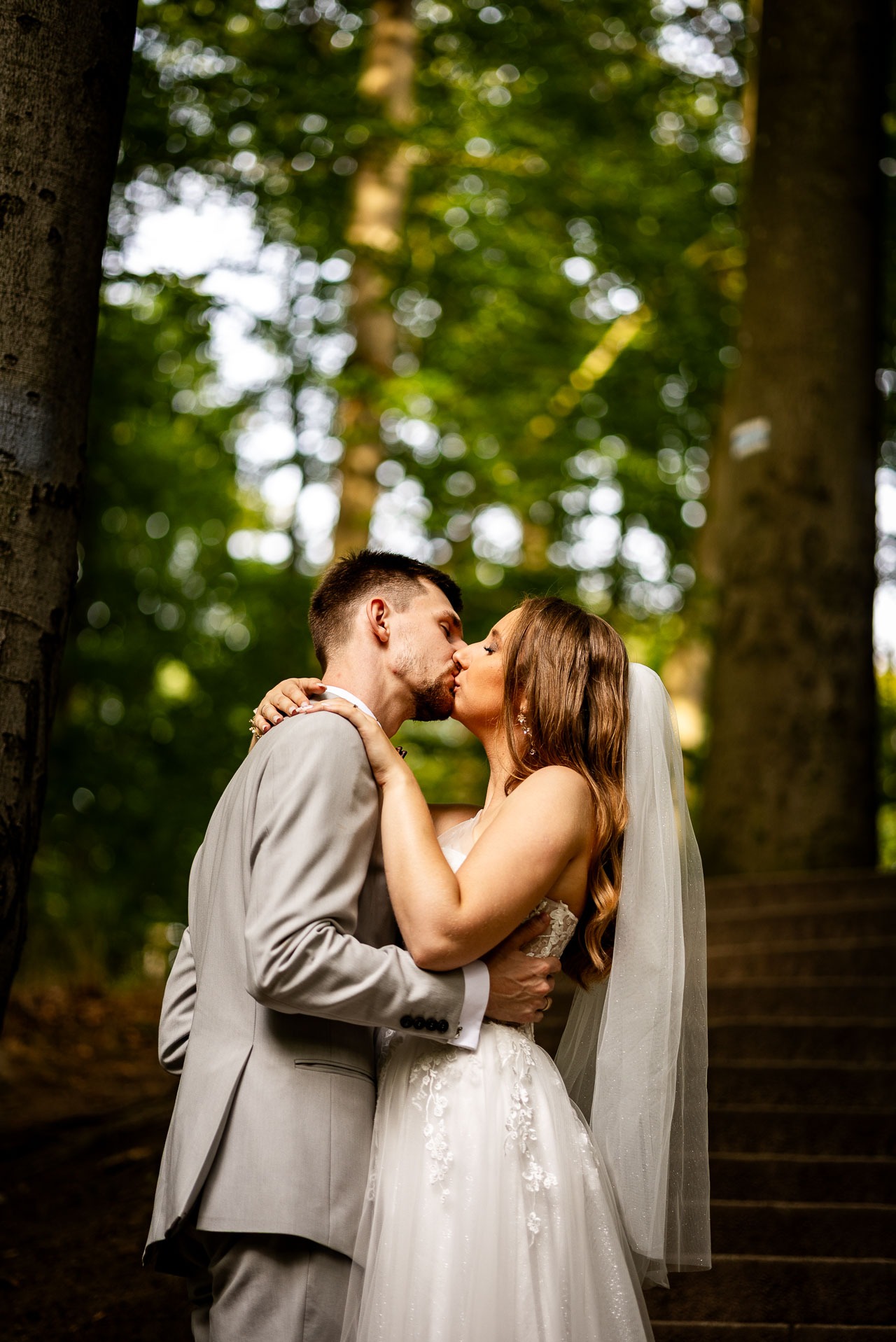 Pocałunek młodej pary w leśnej scenerii. Panna młoda w białej sukni z welonem, pan młody w szarym garniturze. Romantyczna atmosfera, naturalne światło.