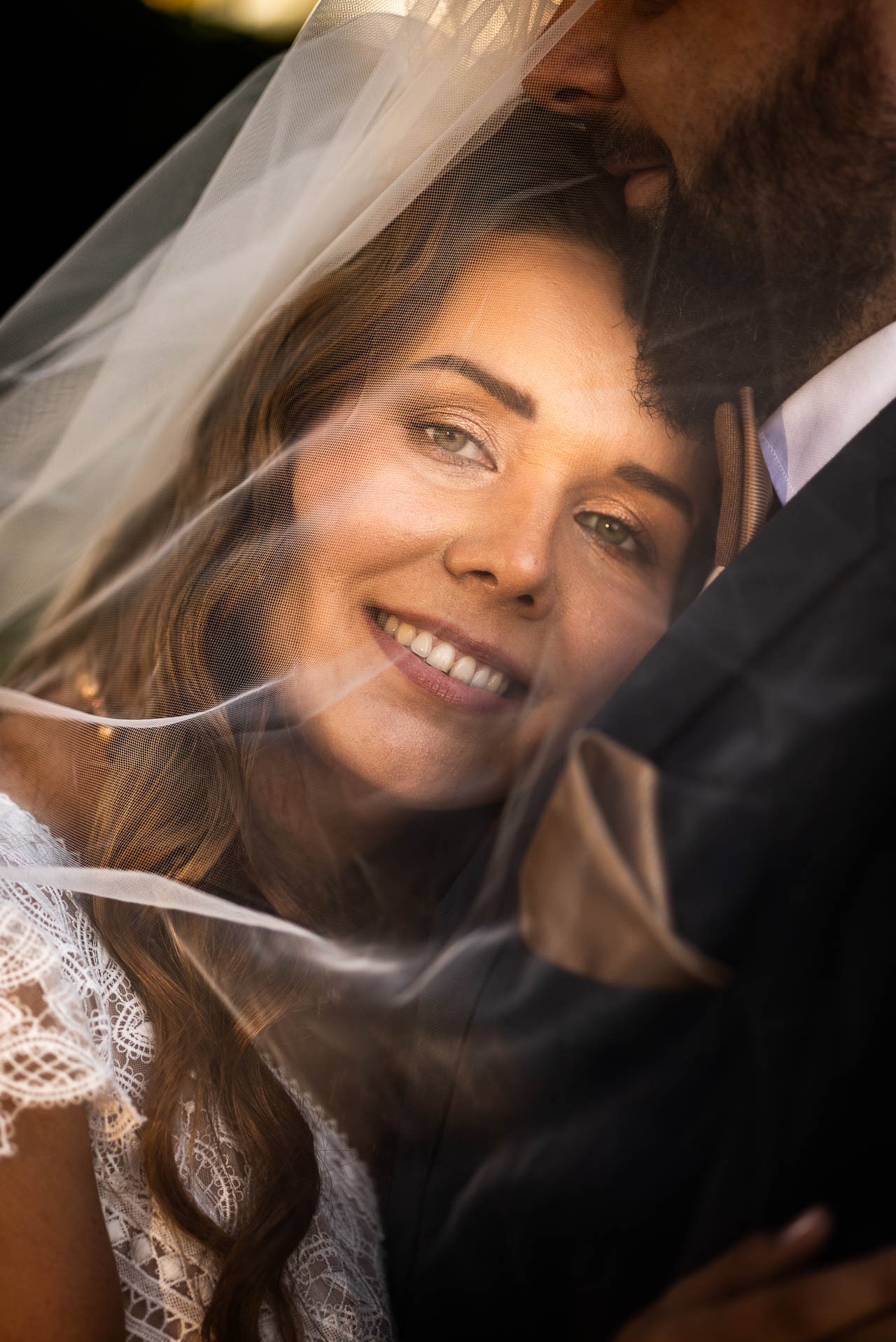 Uśmiechnięta panna młoda pod welonem, wtulona w pana młodego w ciemnym garniturze. Delikatne światło podkreśla makijaż i koronkę sukni.