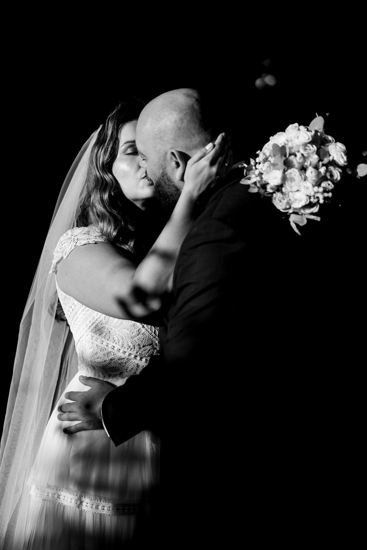 Czarno-białe zdjęcie całującej się pary młodej. Panna młoda w koronkowej sukni i welonie, pan młody w ciemnym garniturze. W tle bukiet ślubny i ciemne tło.