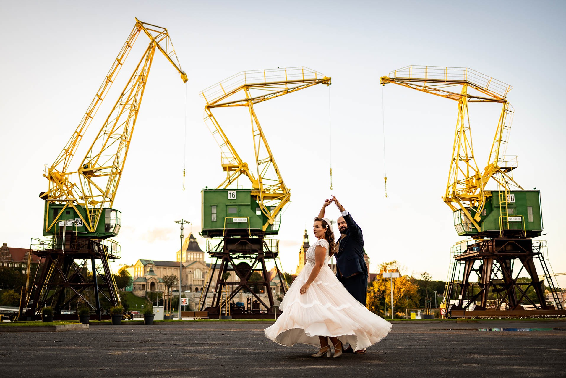 Para młoda tańczy na tle portowych dźwigów w Szczecinie. Panna młoda w sukni wiruje, pan młody w garniturze. W tle budynki i jasne niebo.