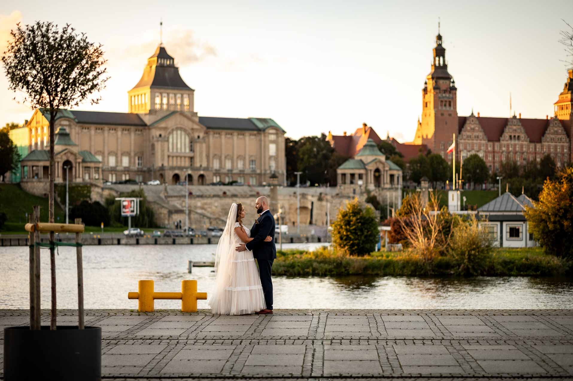 Para młoda w Szczecinie pozuje na tle Odry i Wałów Chrobrego w ciepłym, zachodzącym słońcu. Panna młoda w zwiewnej sukni, pan młody w granatowym garniturze.