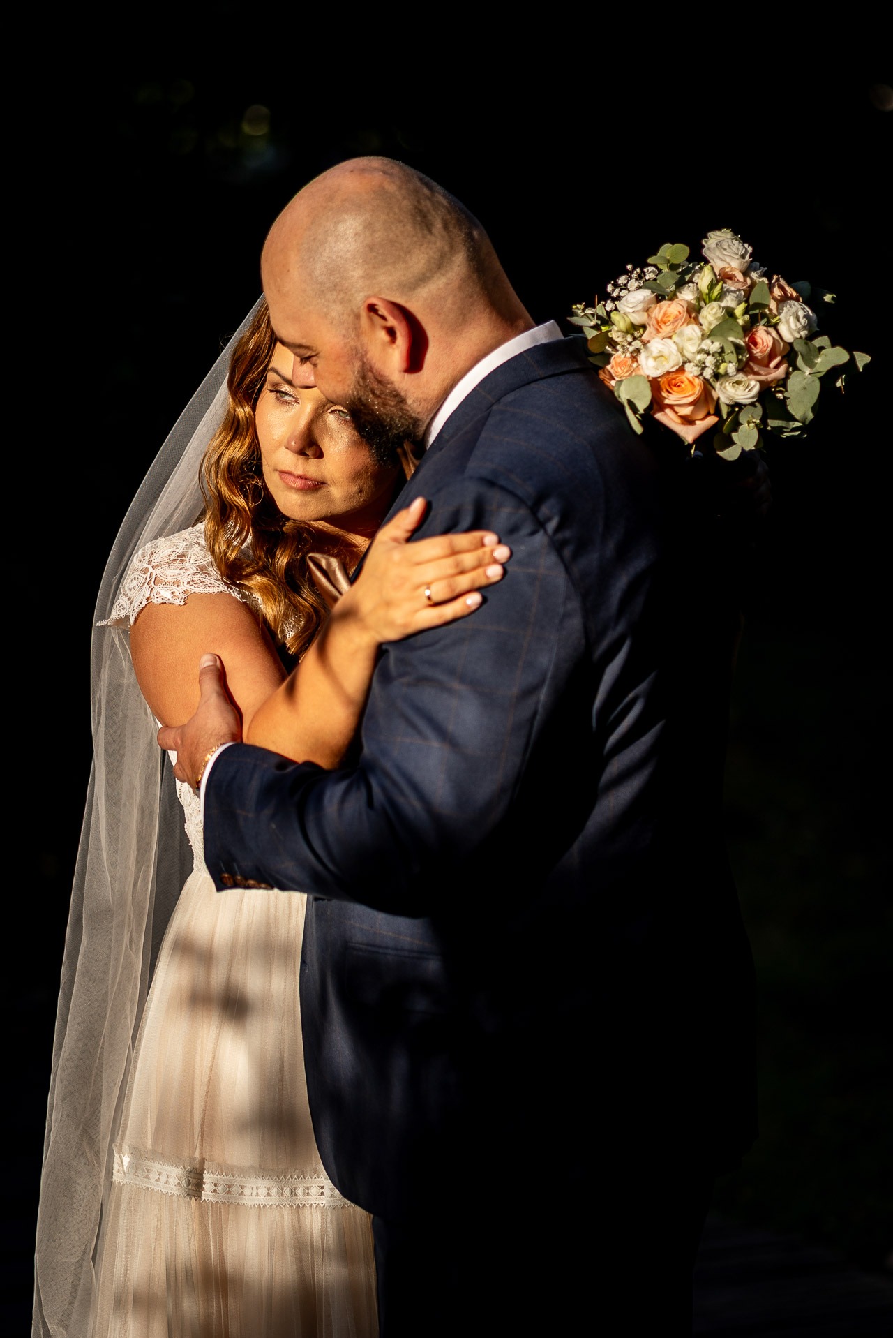 Para młoda w objęciach, panna młoda z bukietem w pastelowych kolorach, oświetlona ciepłym światłem na ciemnym tle, subtelny uśmiech kobiety.