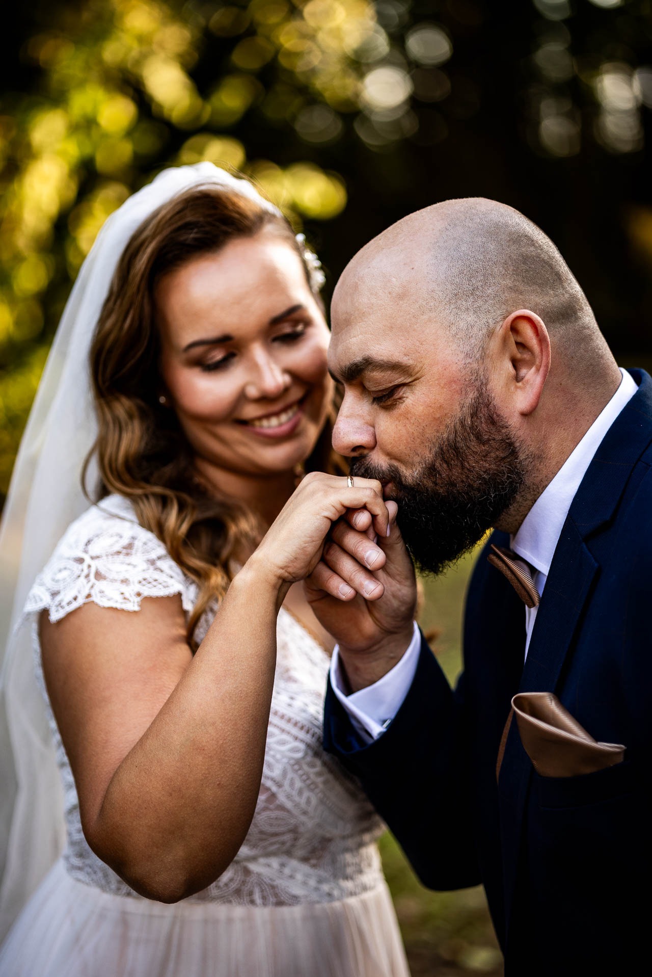 Panna młoda w koronkowej sukni uśmiecha się, gdy pan młody w granatowym garniturze całuje jej dłoń z obrączką na tle zieleni.