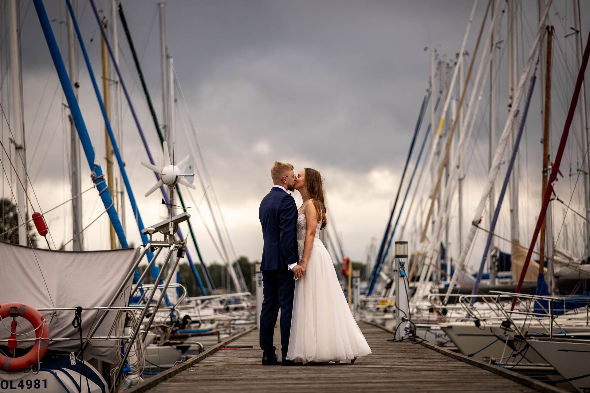 Para młoda całuje się na drewnianym molo, w tle maszty jachtów i pochmurne niebo. Ujęcie z dołu podkreśla wysokość masztów.