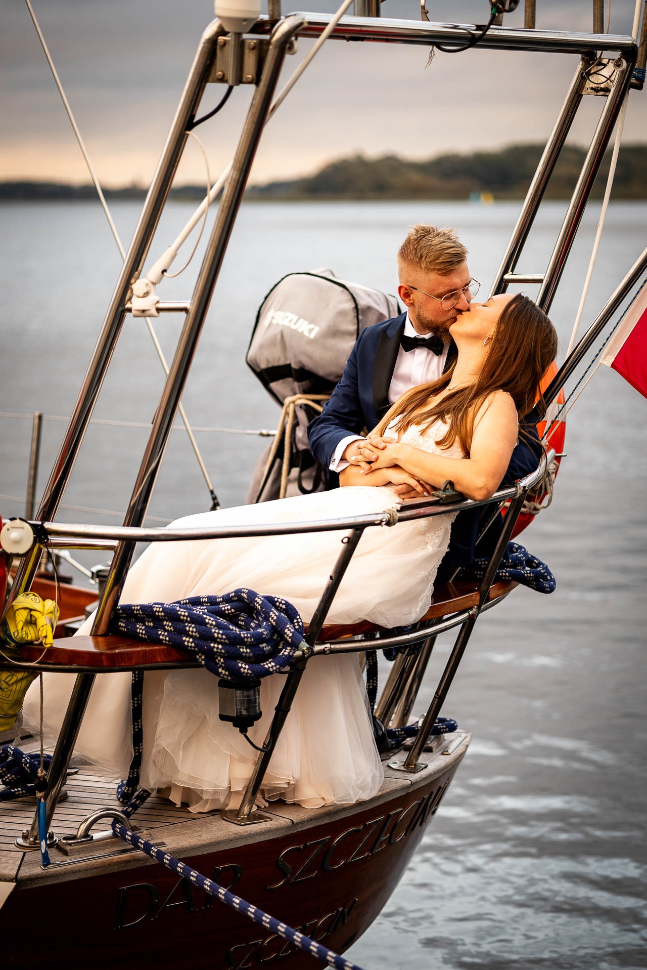 Para młoda w objęciach, pocałunek na tle jachtu z widokiem na jezioro. Pan młody w granatowym smokingu, panna młoda w białej sukni. Romantyczna sesja na wodzie.