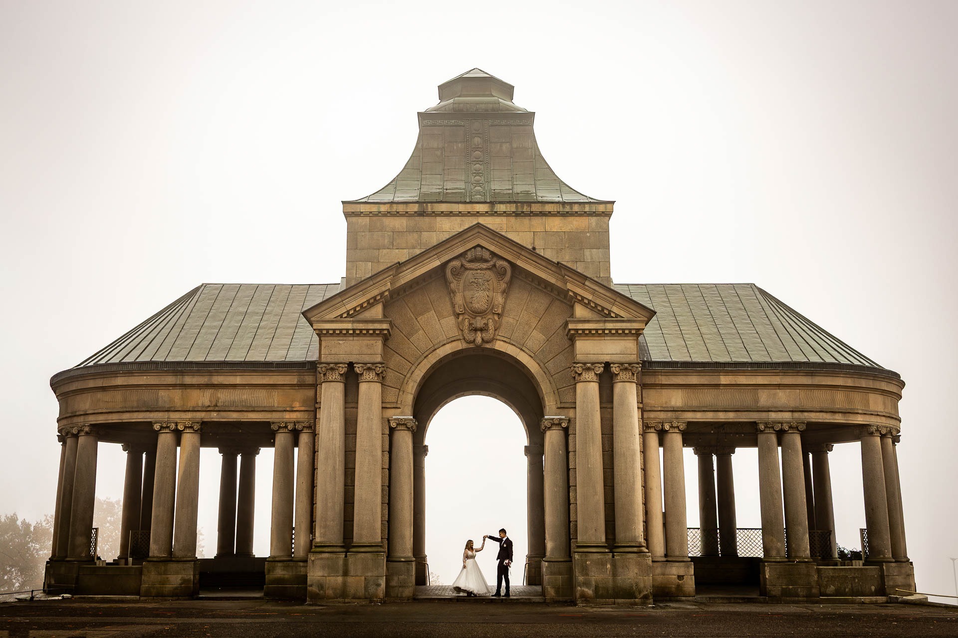 Para młoda tańczy w monumentalnym, kamiennym pawilonie w mglisty dzień. Klasyczna architektura, kolumny i subtelne światło tworzą romantyczną atmosferę.