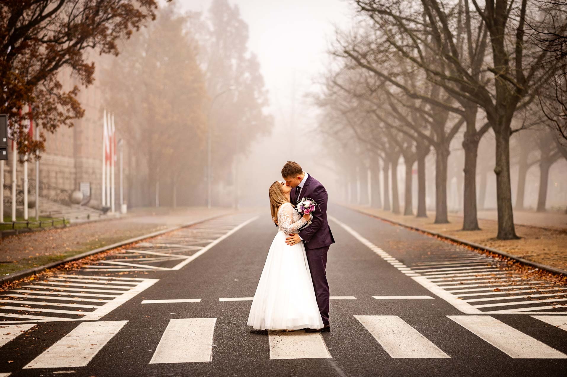 Para młoda całuje się na pasach dla pieszych w mglisty dzień. Panna młoda w białej sukni, pan młody w fioletowym garniturze. W tle drzewa i budynek z flagami.