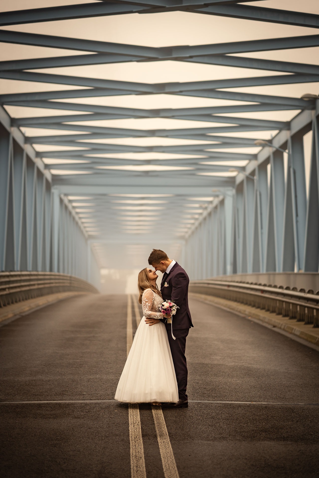 Para młoda w objęciach na moście w mglisty dzień. Panna młoda w koronkowej sukni, pan młody w ciemnym garniturze. Romantyczna sesja w plenerze.