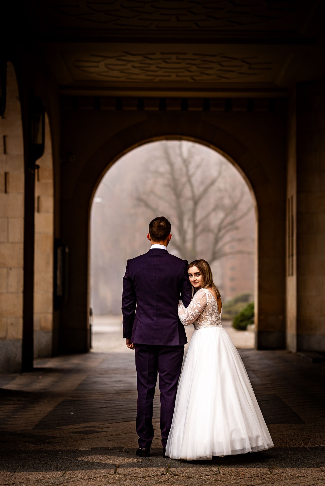 Para młoda w tunelu z łukiem. Panna młoda opiera się o ramię pana młodego, spoglądając w obiektyw. W tle drzewo i budynek w lekkiej mgle.