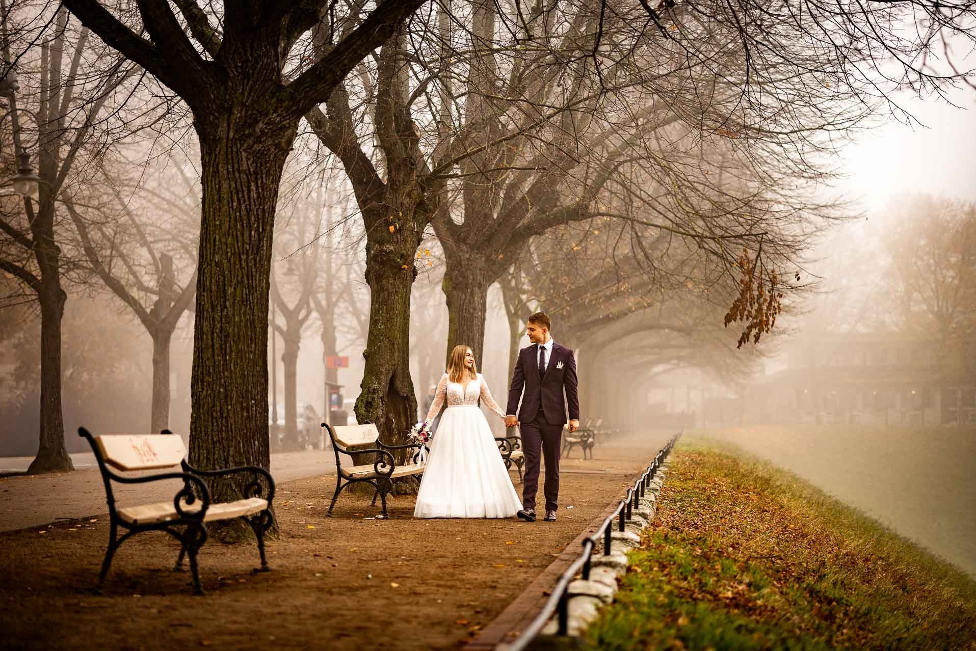 Para młoda spaceruje w parku we mgle, trzymając się za ręce. Panna młoda w białej sukni, pan młody w ciemnym garniturze. W tle drzewa i ławki.