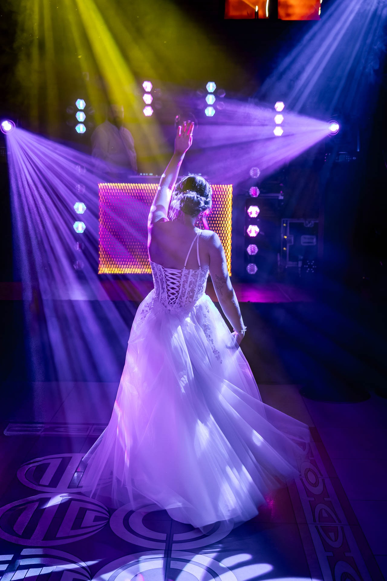 Panna młoda w białej sukni z koronkowym gorsetem, tyłem do obiektywu, tańczy na parkiecie w fioletowych światłach dyskoteki. DJ w tle.