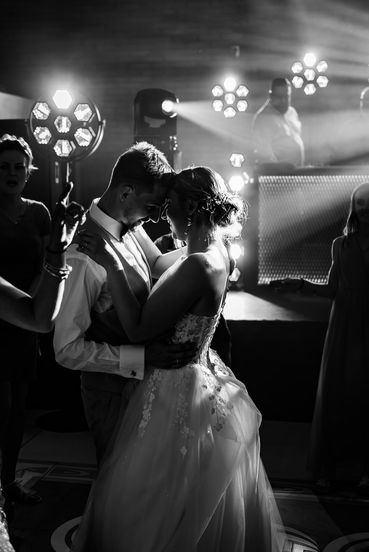 Czarno-białe zdjęcie tańczącej pary młodej w Szczecinie, oświetlonej reflektorami, z gośćmi weselnymi w tle. Suknia panny młodej zdobiona kwiatowym wzorem.