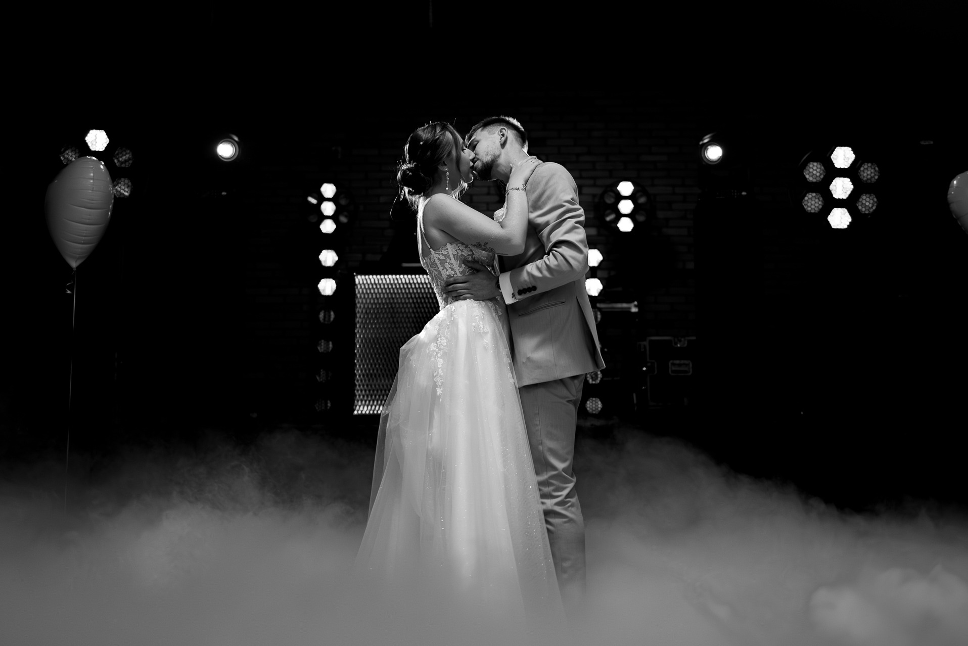 Czarno-białe zdjęcie całującej się pary młodej na tle świateł i dymu. Panna młoda w sukni z koronkową górą, pan młody w garniturze. Romantyczna atmosfera.