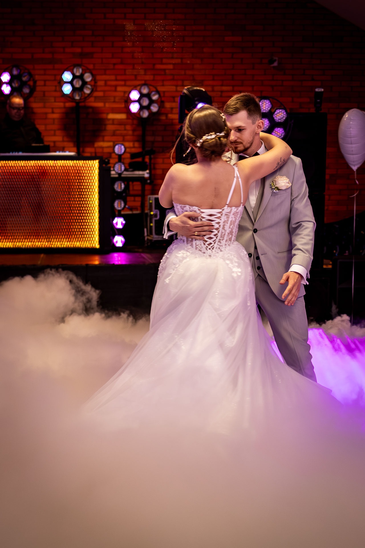 Para młoda w tańcu na tle ceglanej ściany z oświetleniem scenicznym i efektem mgły. Suknia z koronkowym gorsetem, garnitur w odcieniach szarości. Romantyczna atmosfera.